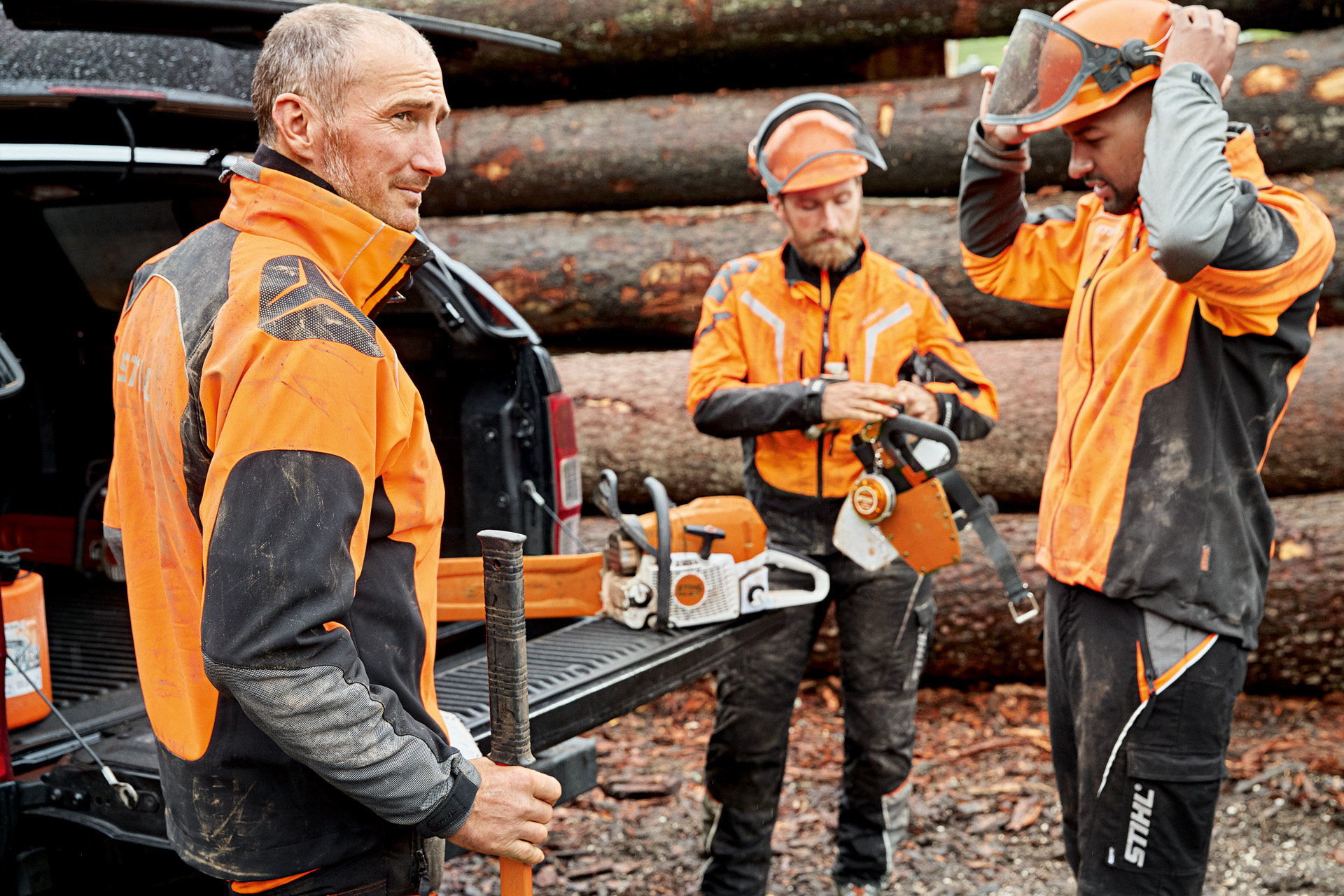 Trois hommes en vêtements de protection STIHL devant un tas de bois se préparent à travailler avec une tronçonneuse STIHL